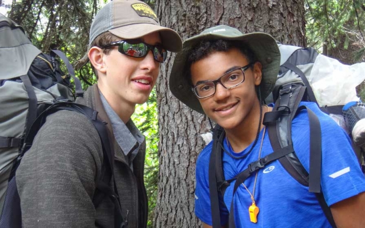 Two students wearing backpacks smile at the camera in front of a tree. 